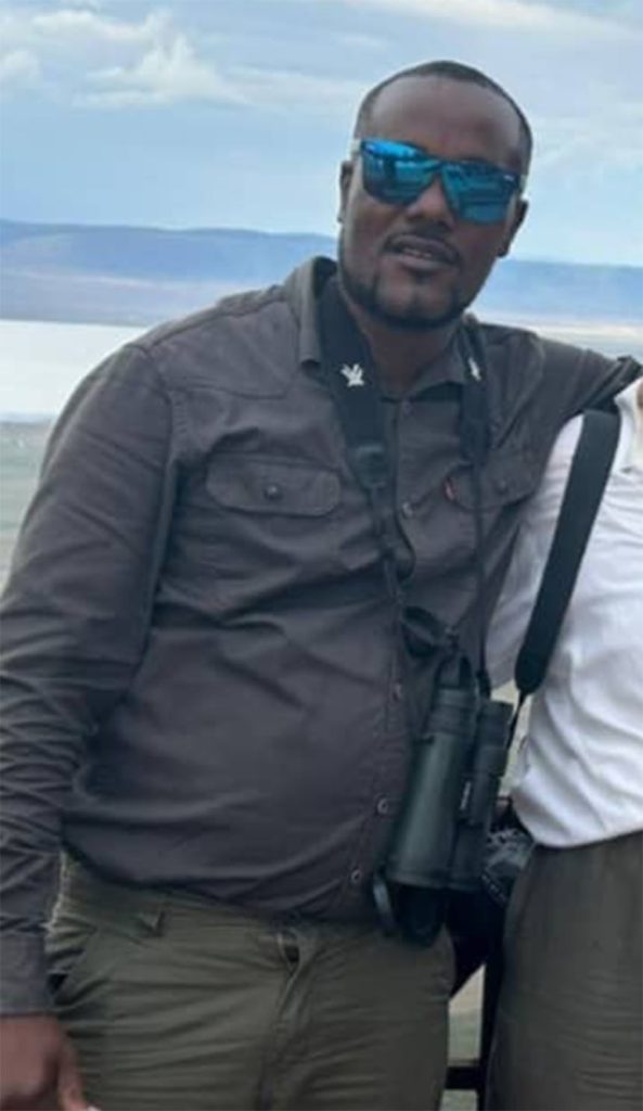 Man in a gray button-front shirt and olive pants, wearing blue mirrored sunglasses, outdoors by a lake with hills in the background.