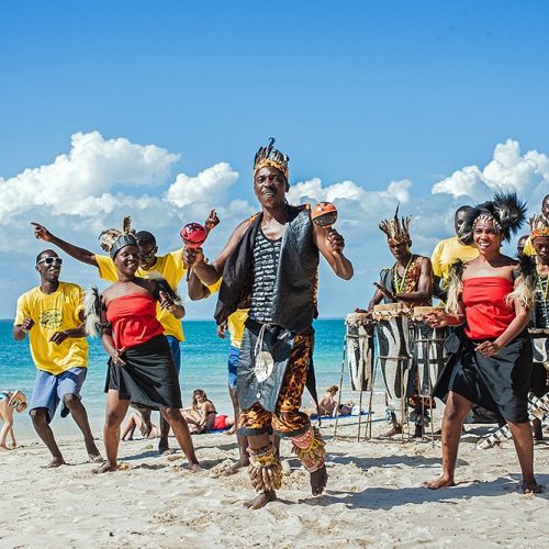 Dancers perform on a sunny beach with a drummer group in yellow shirts watching, ocean in the background.