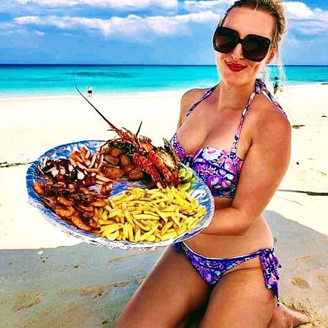 Woman in a purple bikini and sunglasses smiles while presenting a large plate of seafood and fries on a sunny beach with turquoise water behind her,