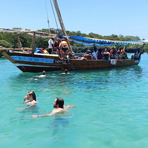 Group of people on a wooden sailboat with a blue canopy, anchored in turquoise water as two swimmers approach from nearby.